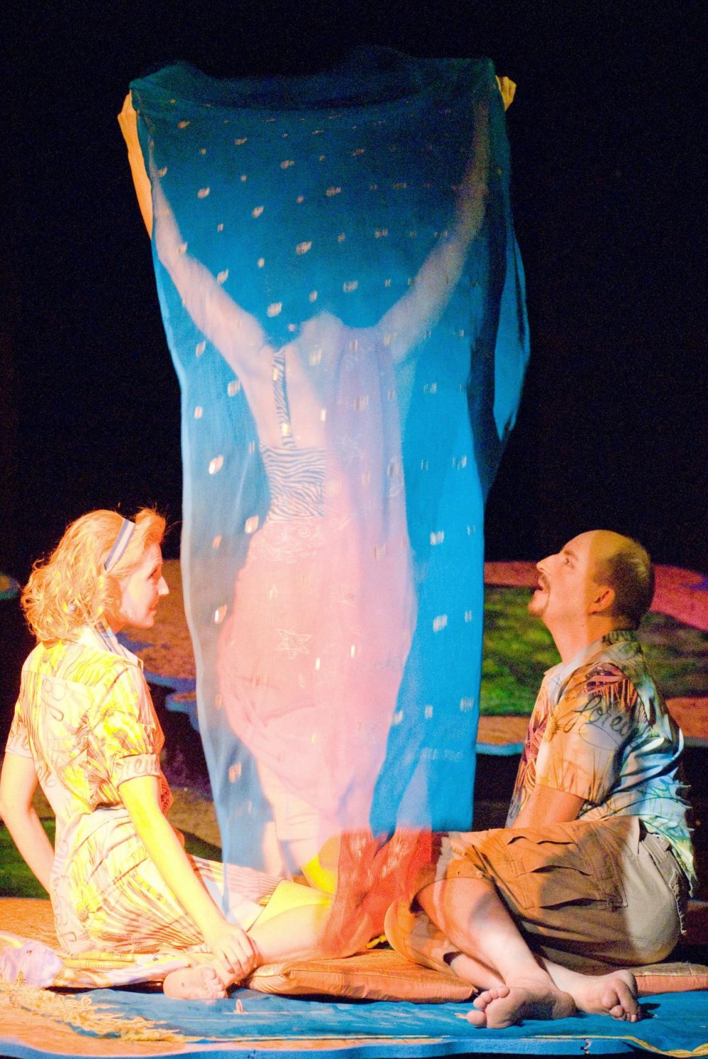 Blonde Frau in hellem Kleid und Mann mit buntem Hemd und brauner kurzer Hose sitzen auf der Bühne. Zwischen Ihnen eine Person, die ein blaues durchsichtiges Tuch vom Boden nach oben hält. 