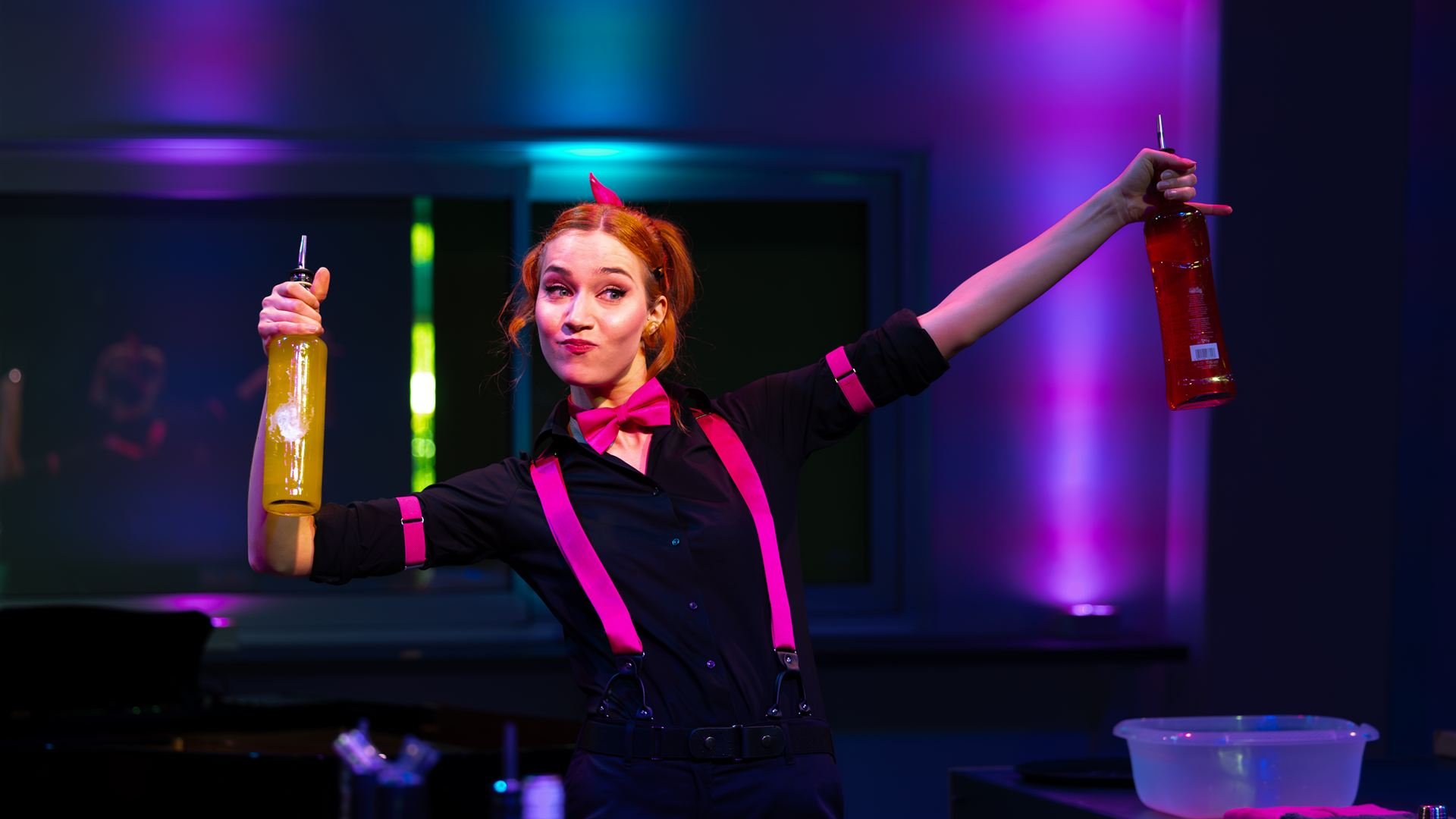 Carmen hält tanzend zwei Flaschen mit bunten Getränken in die Luft, während sie hinter dem Tresen steht.