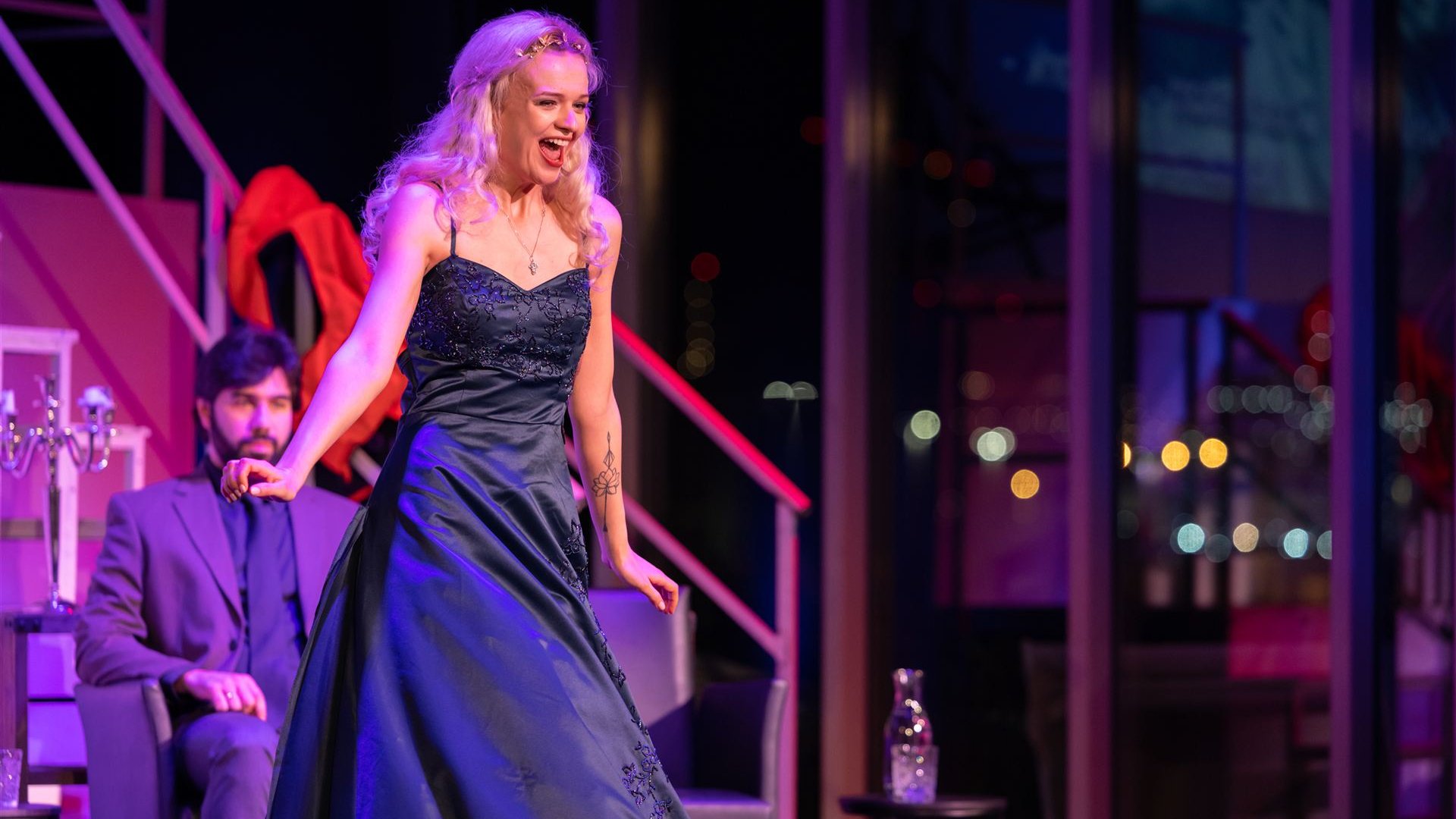 Eine Sängerin steht bei einem Opern-Slam auf der Bühne und singt. Im Hintergrund sitzt ein Sänger und schaut ihr zu.